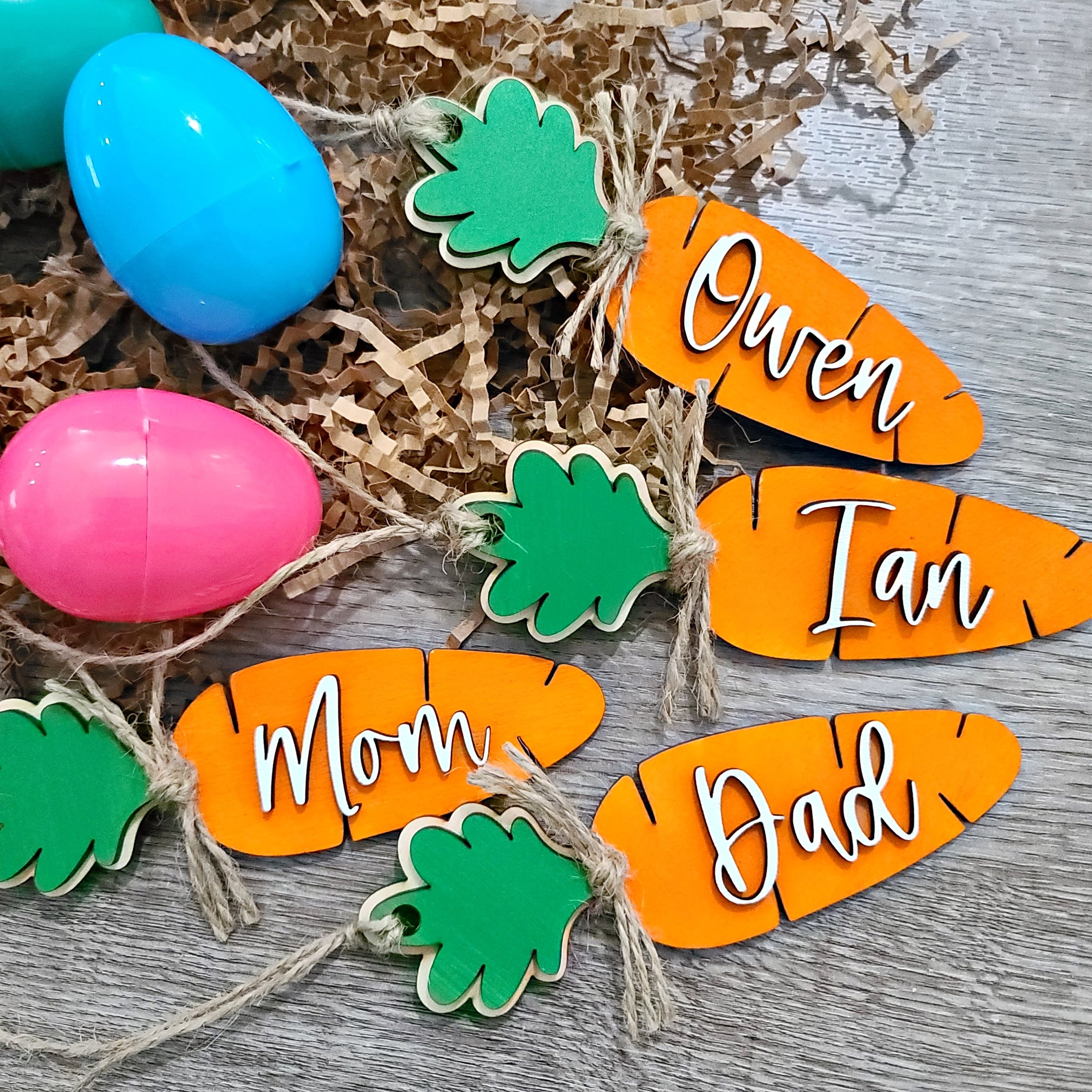 4 orange wooden cutout carrots with a 3D green leaf. A white script 3D name is attached to the long side of the carrot. Twine is attached as a hanger to the green leaf and a twine knot embellishment is at the neck of the carrot.
