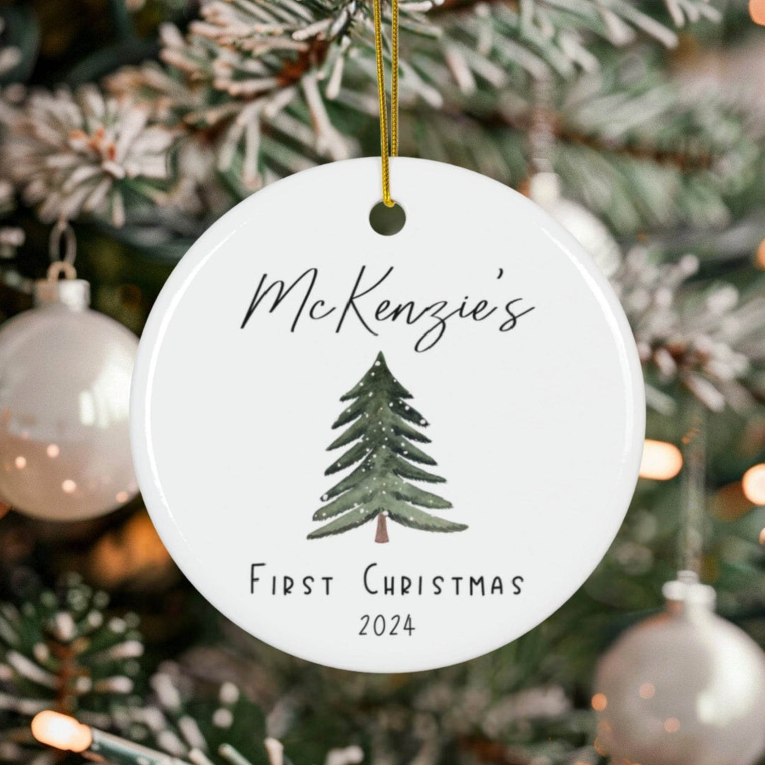 Round white ceramic Christmas ornament with tree design and text 'McKenzie's First Christmas 2024' on a decorated tree.