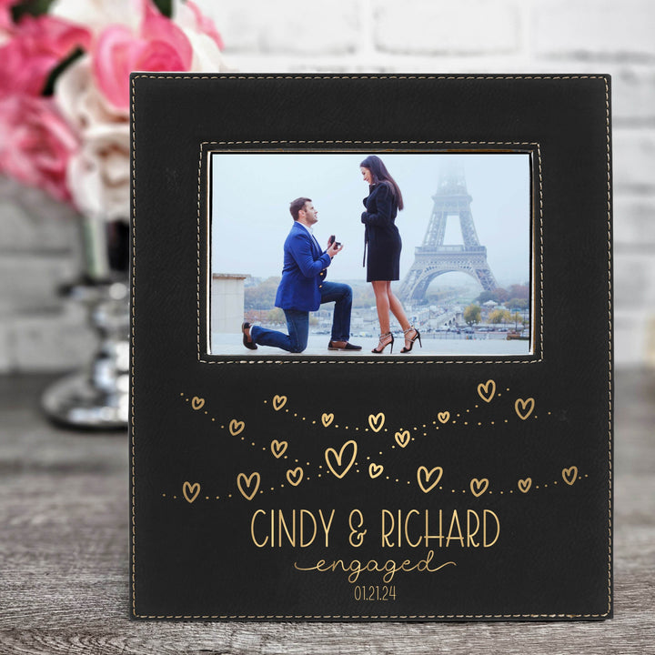 Personalized black vegan leather engagement photo frame with engraved names and text 'Cindy & Richard engaged 01.21.24' in metallic gold on a wooden surface.