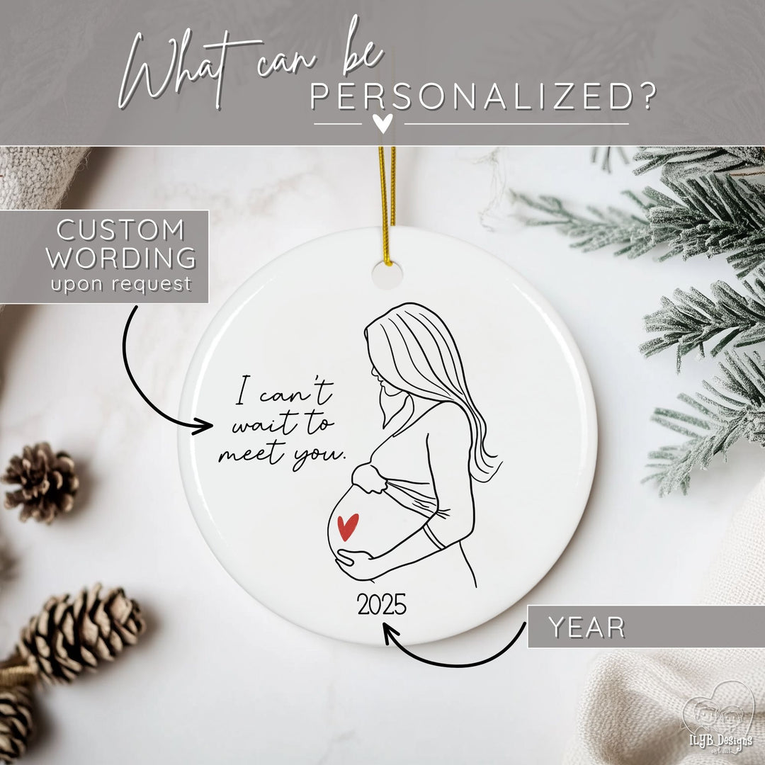 Graphic showing what part of the ornament can be personalized. Arrows point to the year and custom wording where "I can't wait to meet you" is written. Image is of a round white ceramic pregnancy Christmas ornament featuring a black and white line drawing of a pregnant woman holding her belly with a red heart accent on her baby bump. The sweet black script says “I can’t wait to meet you” and the year on the bottom make it a meaningful pregnancy ornament and keepsake.