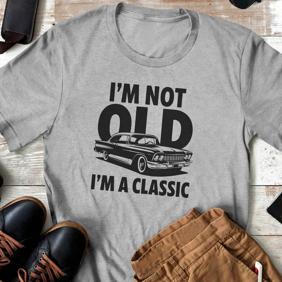 Gray t-shirt with a graphic and text 'I'm not old, I'm a classic' on a wooden surface with shoes and pants.