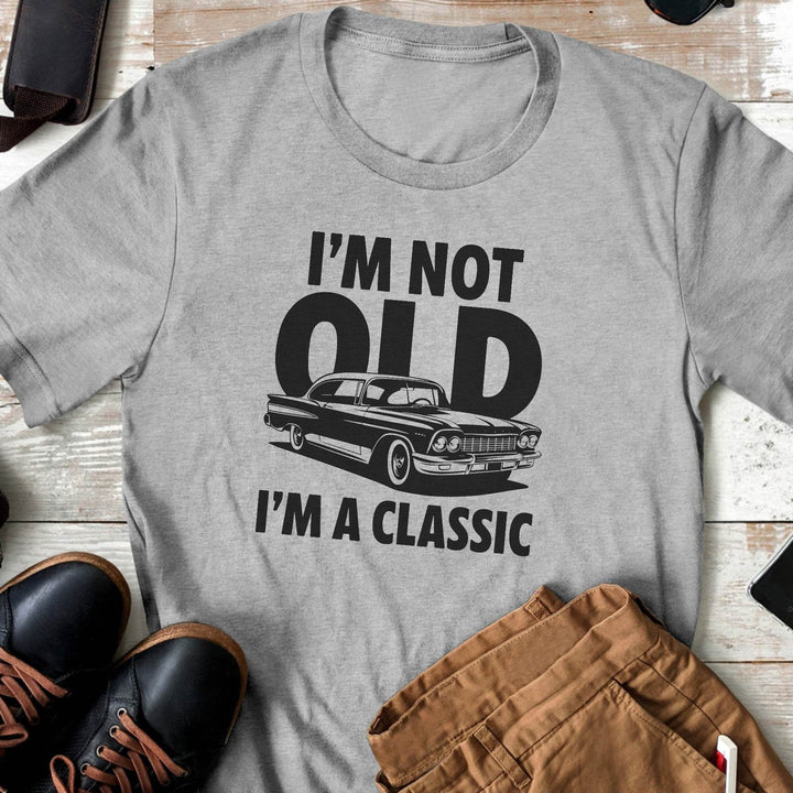 Gray t-shirt with a graphic and text 'I'm not old, I'm a classic' on a wooden surface with shoes and pants.