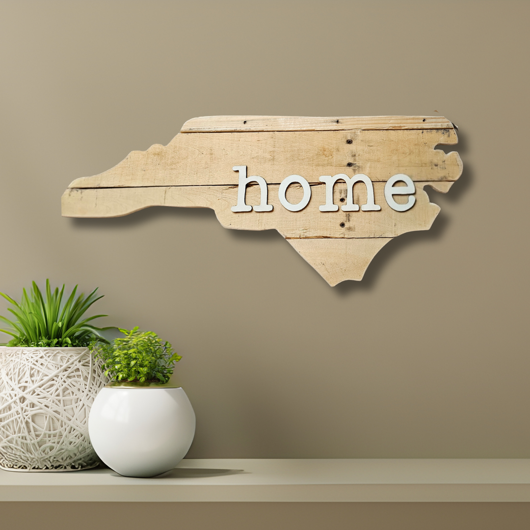 Wood pallet wall art shaped like state of North Carolina with 3D 'home' text, placed on a shelf with plants against a beige wall.
