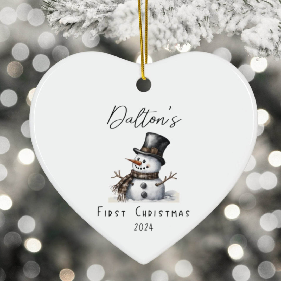 Heart-shaped ornament with a snowman illustration and text 'Dalton's First Christmas 2024' against a blurred Christmas tree background.