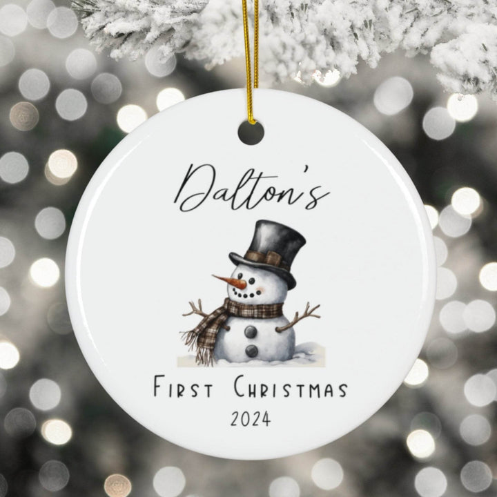 Round Christmas ornament with a snowman and text 'Dalton's First Christmas 2024' against a blurred Christmas tree background.