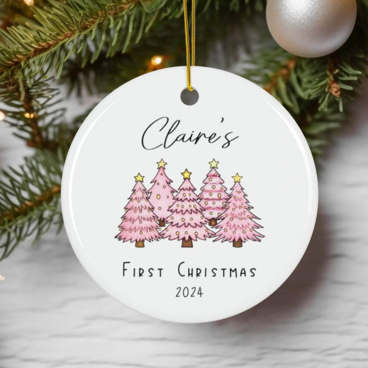 Round white Christmas ornament with pink trees and 'Claire's First Christmas 2024' text, hanging on a tree.