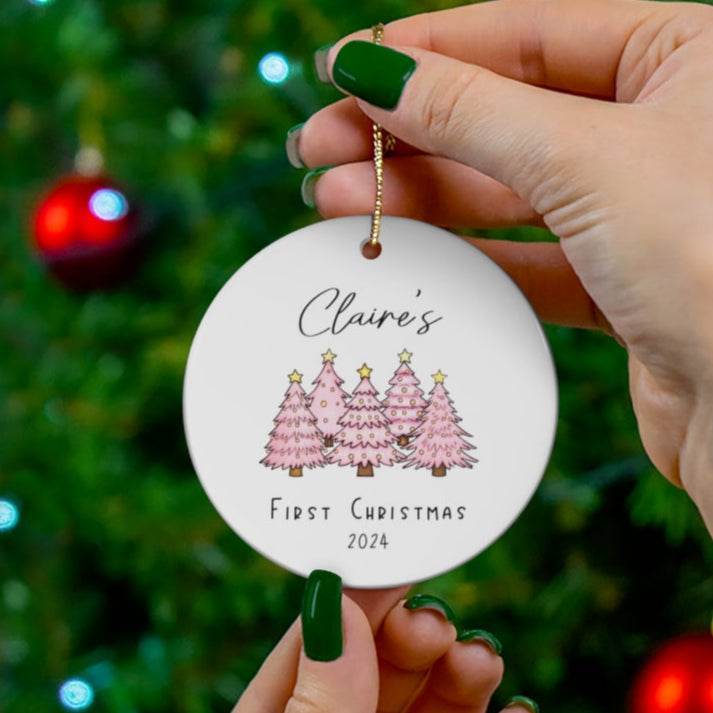 Round Christmas ornament with pink trees and 'Claire's First Christmas 2024' text, held in front of a decorated tree.