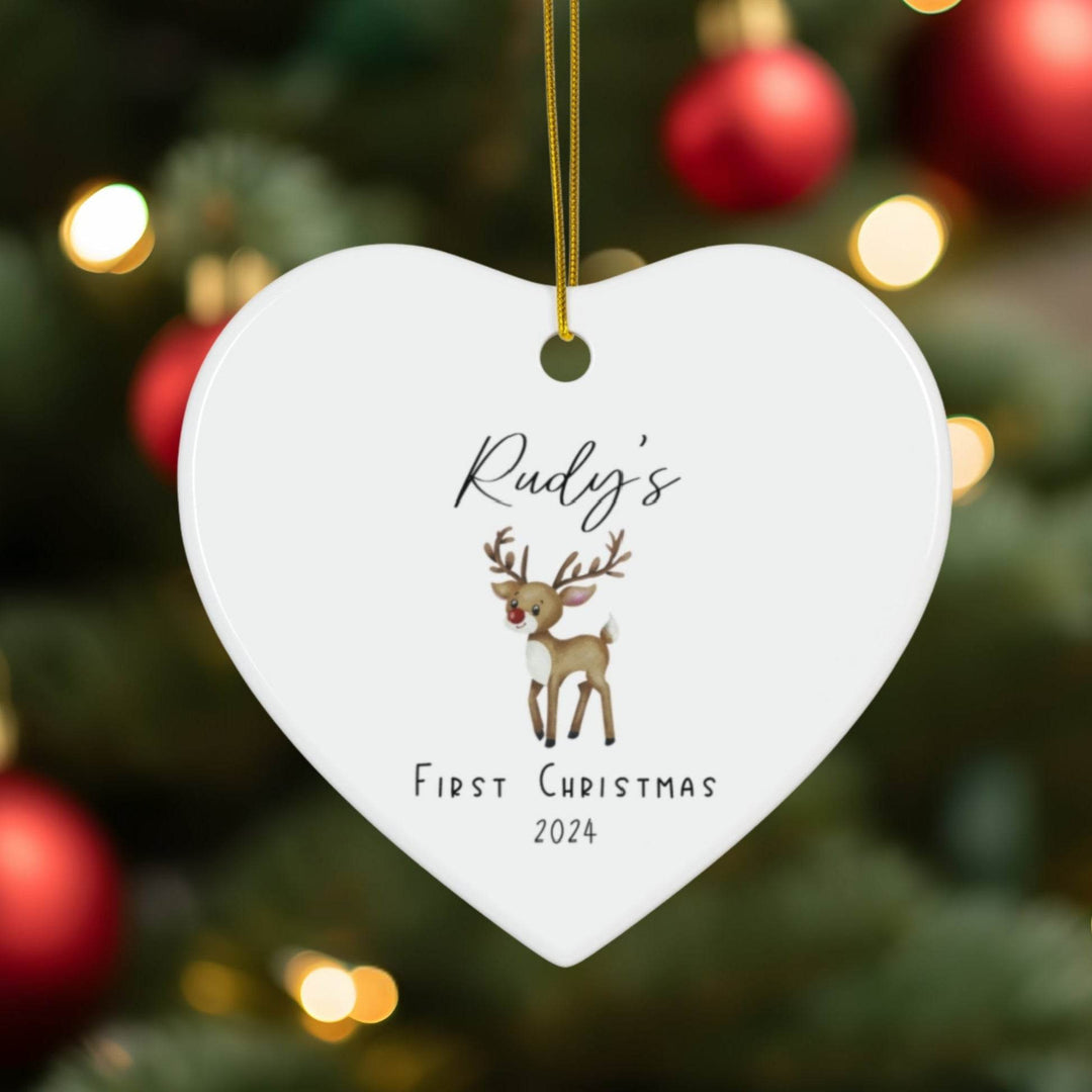 White heart-shaped Christmas ornament with a watercolor red-nosed reindeer illustration and text 'Rudy's First Christmas 2024' against a Christmas tree background.