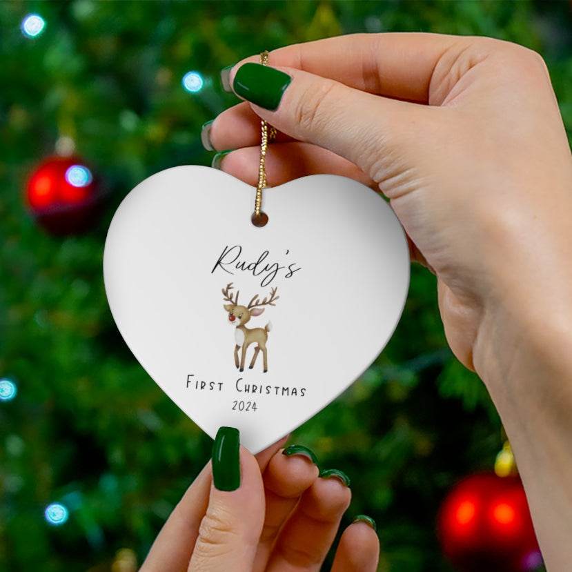 Heart-shaped white ceramic Christmas ornament with reindeer design and text 'Rudy's First Christmas' with year, held in front of a Christmas tree.