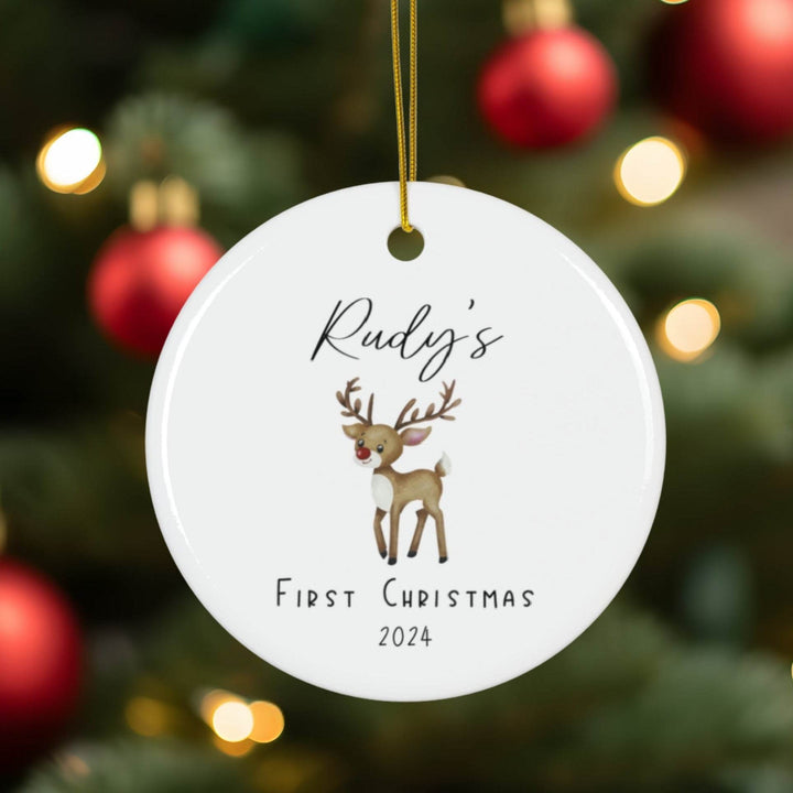 Round white ceramic Christmas ornament with watercolor reindeer illustration and text 'Rudy's First Christmas 2024' against a festive background.