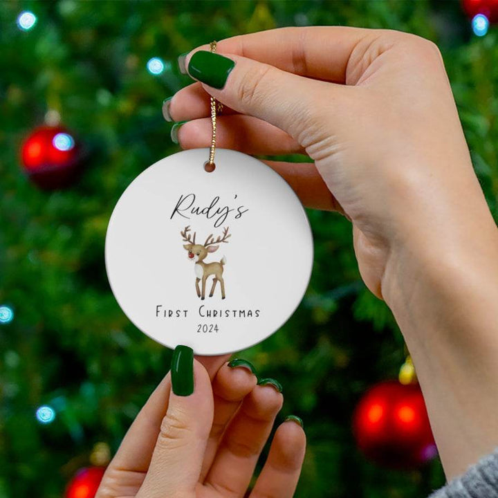 Hand holding a white ceramic round Christmas ornament with a reindeer design and text 'Rudy's First Christmas' and year in front of a decorated tree.