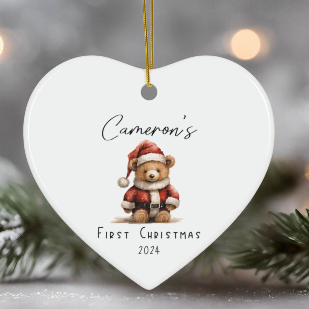 Heart-shaped ornament with a teddy bear in a Santa suit and text 'Cameron's First Christmas 2024' against a blurred Christmas tree background.