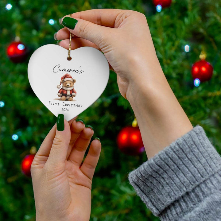 Heart-shaped white ceramic ornament with a teddy bear design held by hands against a Christmas tree background.