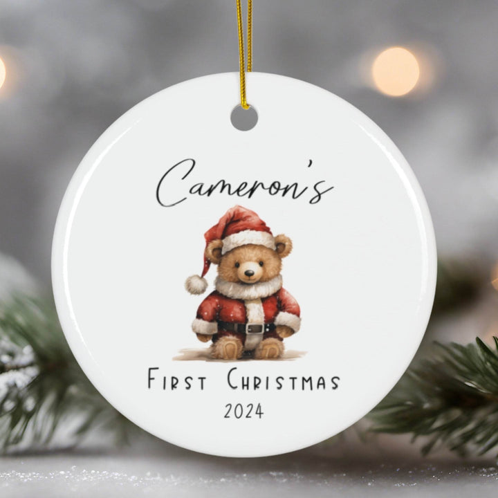 Round Christmas ornament with a teddy bear in a Santa outfit and text 'Cameron's First Christmas 2024' against a blurred Christmas tree background.