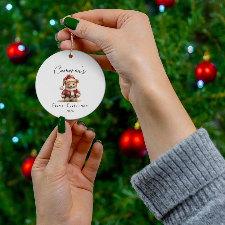 Decorative round ceramic Christmas ornament with a teddy bear and text that says 'Cameron's First Christmas' with the year, held by hands against a Christmas tree background