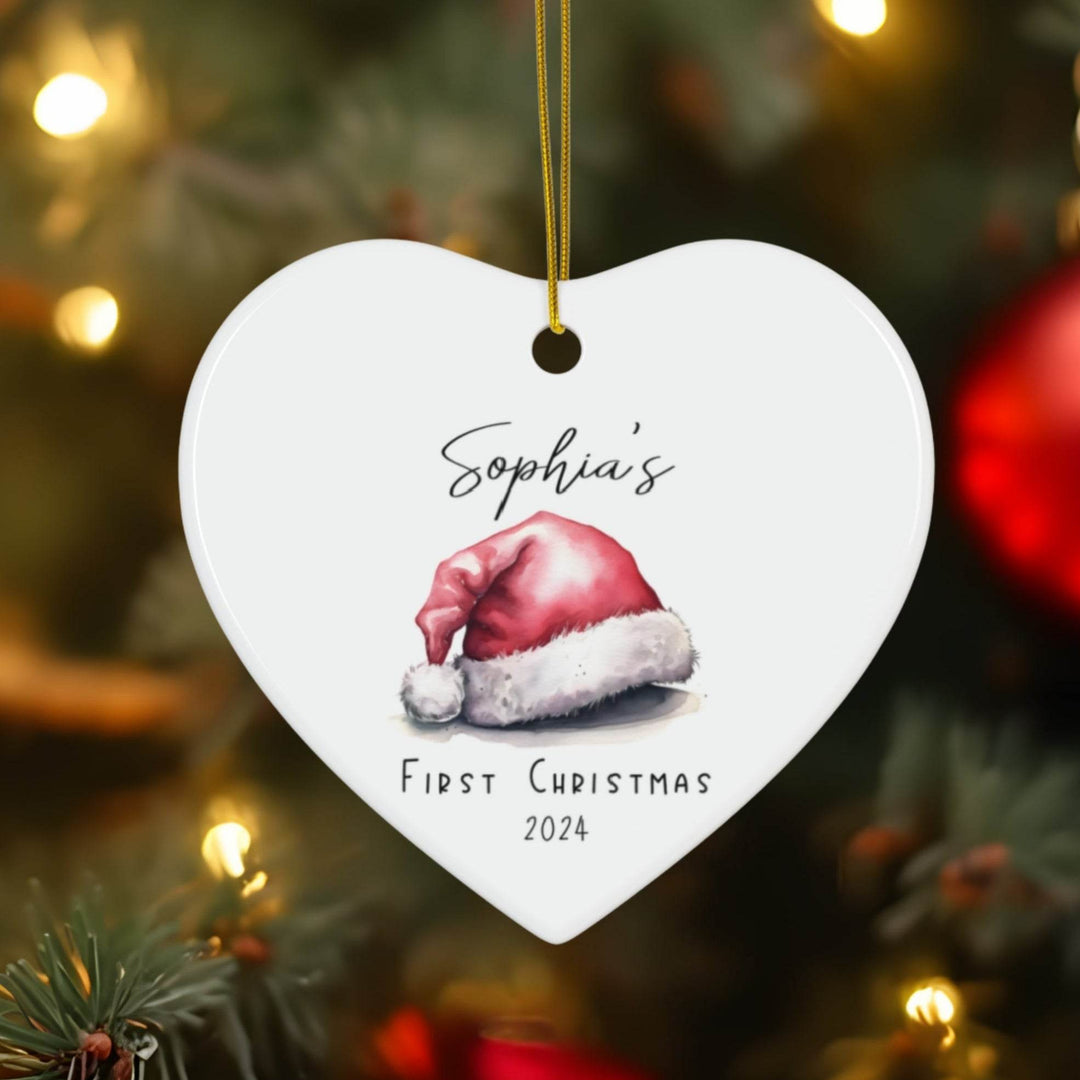 Heart-shaped white ceramic Christmas ornament with Santa hat illustration and text 'Sophia's First Christmas 2024' on a blurred Christmas tree background.