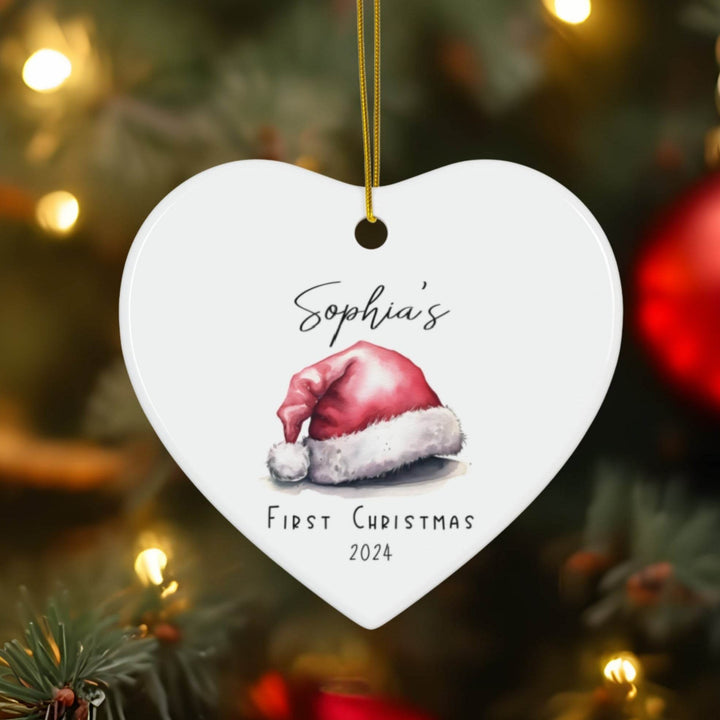 Heart-shaped white ceramic Christmas ornament with Santa hat illustration and text 'Sophia's First Christmas 2024' on a blurred Christmas tree background.