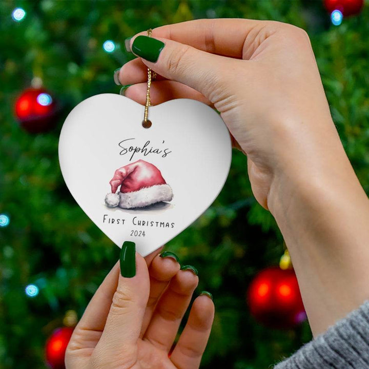 Heart-shaped white ceramic Christmas ornament with 'Sophia's First Christmas 2024' text and Santa hat illustration, held against a Christmas tree background.