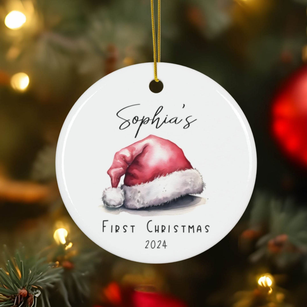 Personalized white ceramic Christmas ornament with watercolor Santa hat and 'Sophia's First Christmas 2024' text on a blurred Christmas tree background.