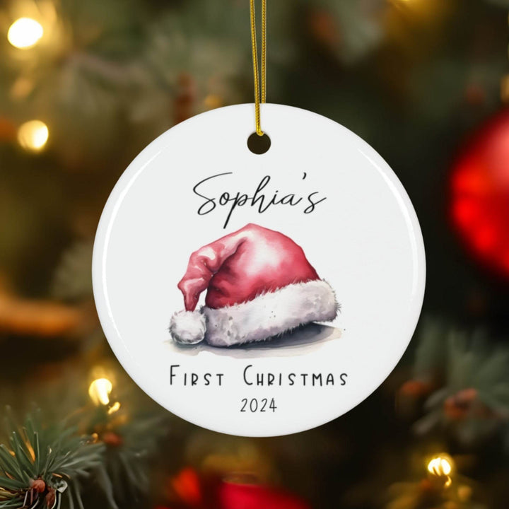 Personalized white ceramic Christmas ornament with watercolor Santa hat and 'Sophia's First Christmas 2024' text on a blurred Christmas tree background.