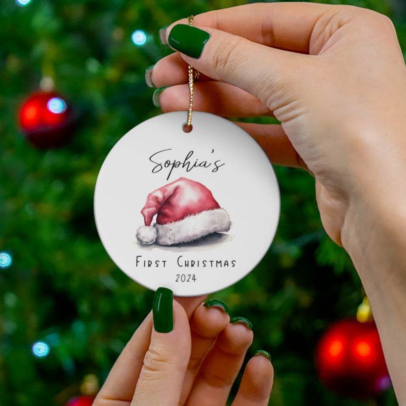 Round white ceramic Christmas ornament with Santa hat design and text 'Sophia's First Christmas 2024' held in front of a Christmas tree.