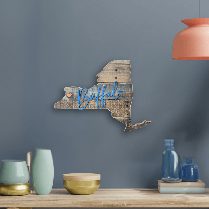 Wooden wall decoration of New York state silhouette cut from pallet wood with blue 3D script 'Buffalo' on a blue wall.