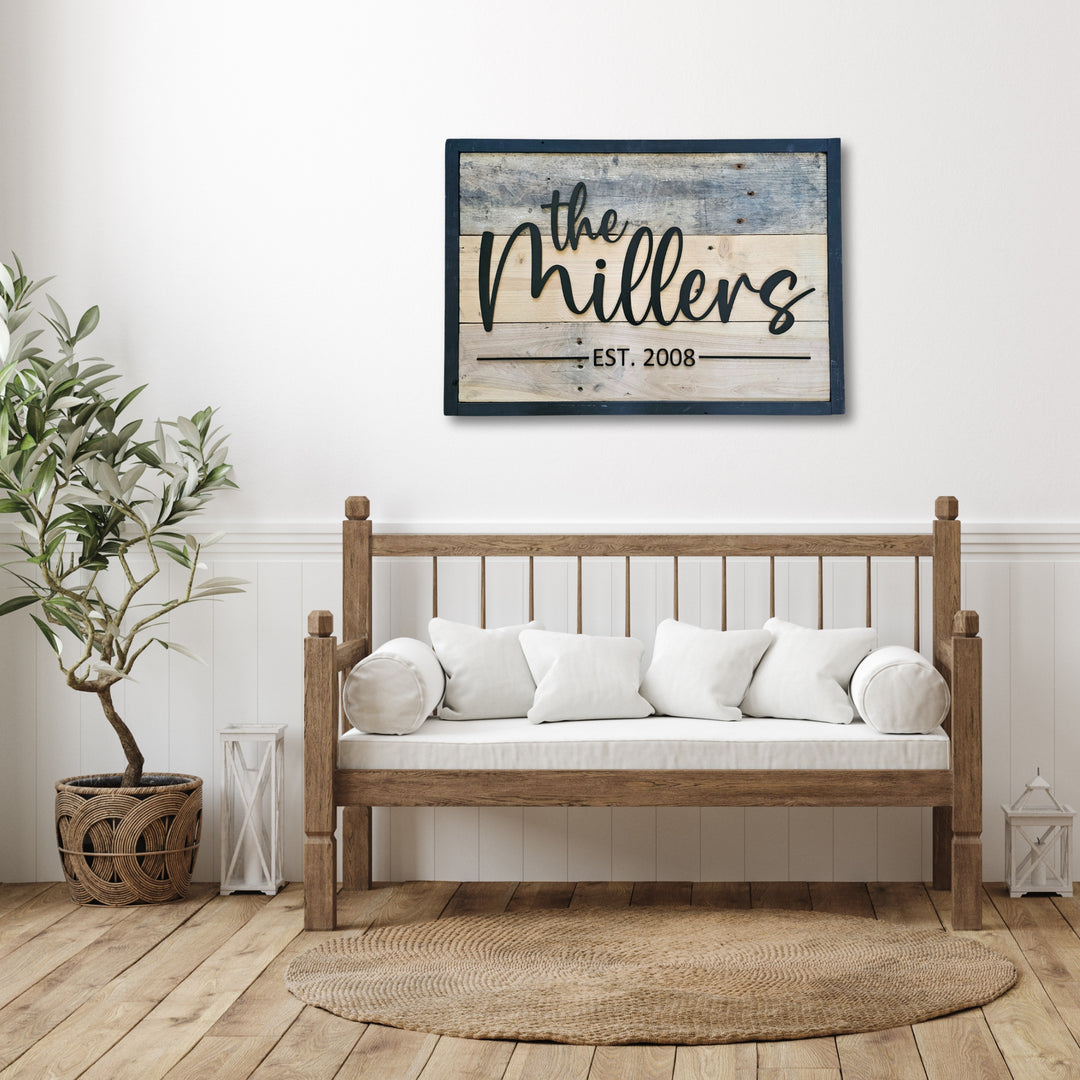 Decorative pallet wall sign with black frame and 'the Millers est. 2008' on a white wall above wooden bench with white cushions.