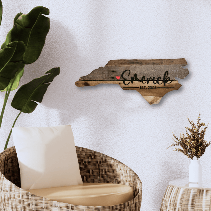 Wooden North Carolina state-shaped pallet sign with 'Emerick' and a heart on a white wall above a wicker chair.