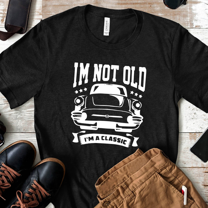 Black t-shirt with 'I'm not old, I'm a classic' graphic on a wooden surface with shoes and shorts.