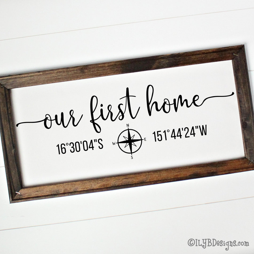 Decorative canvas sign in a brown frame with 'our first home' in black script text along with coordinates and a compass on a white canvas background.