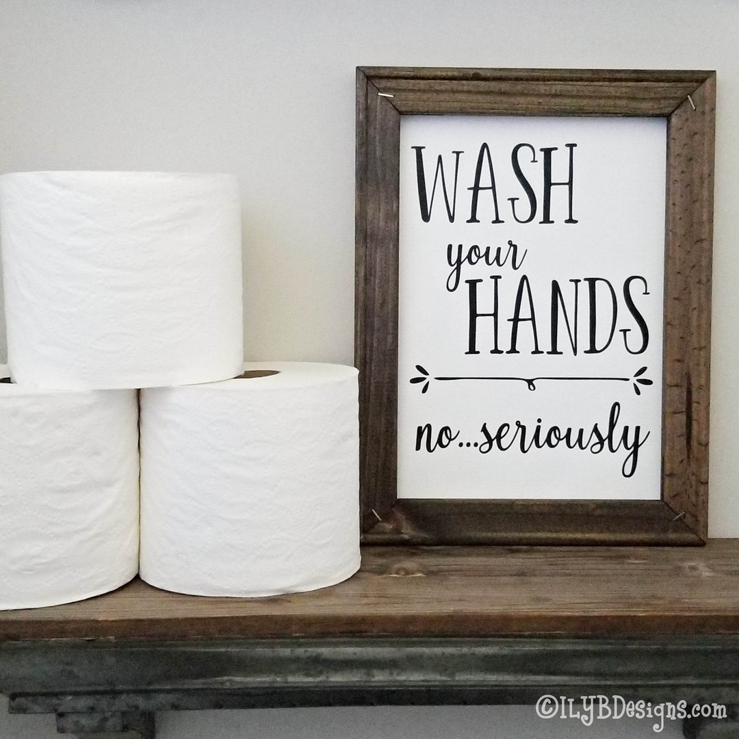 Framed canvas sign with 'Wash your hands no...seriously' black text on a white canvas background with a brown frame sitting on a bathroom shelf next to 3 rolls of toilet paper.
