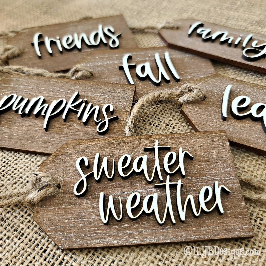 a collection of fall script words on a wood cutout tag with twine to be used as fall napkin holder