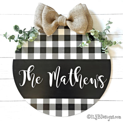 Round door hanger with black and white buffalo plaid on the top and bottom. In the middle is a wide black stripe with a family name in white script. There is a burlap bow and greenery at the top of the sign.