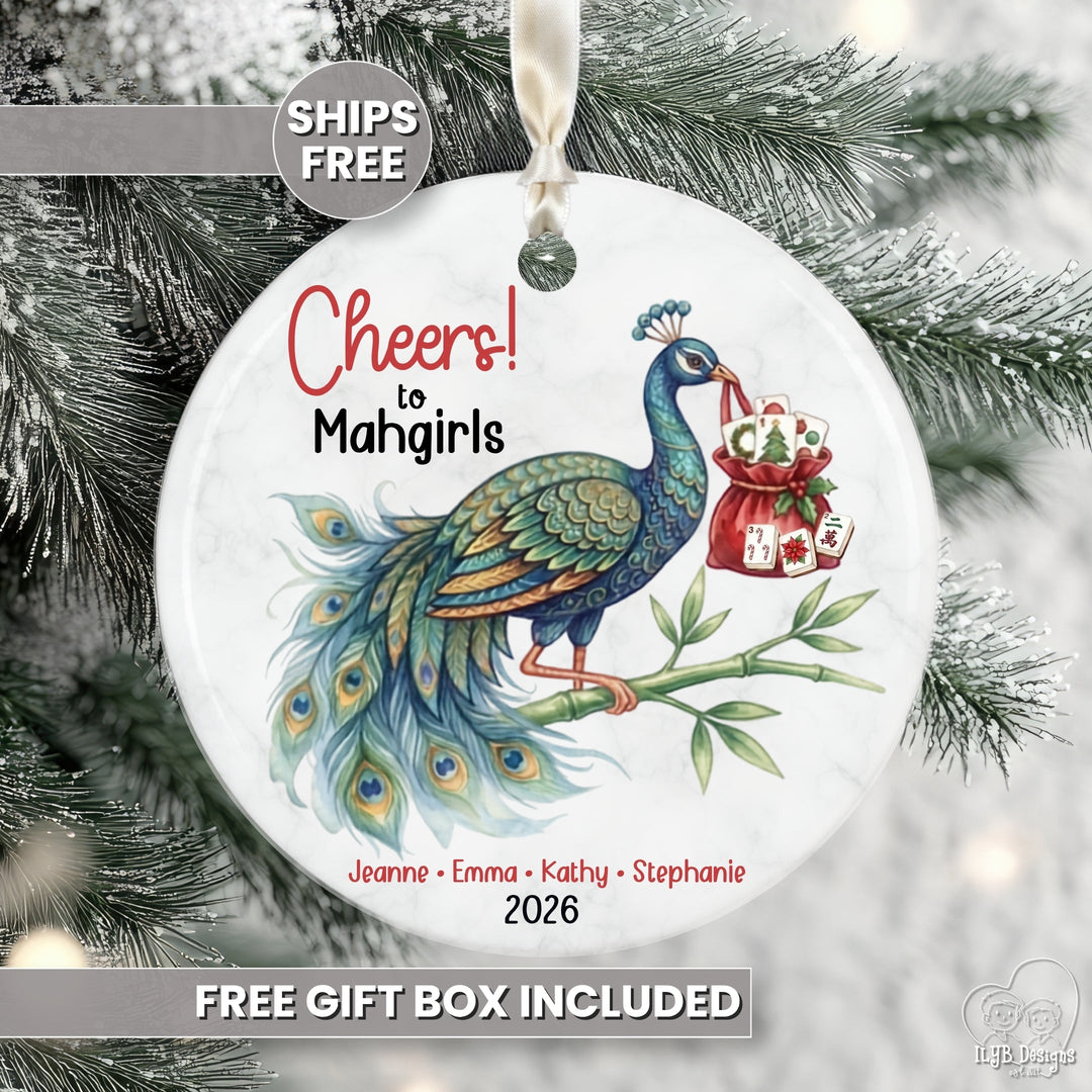 Round white Christmas ornament featuring a colorful peacock illustration and the text 'Cheers to Mahgirls' with names and the year 2026, hanging against a snowy tree background. Promotional text overlays say 'SHIPS FREE' and 'FREE GIFT BOX INCLUDED'.