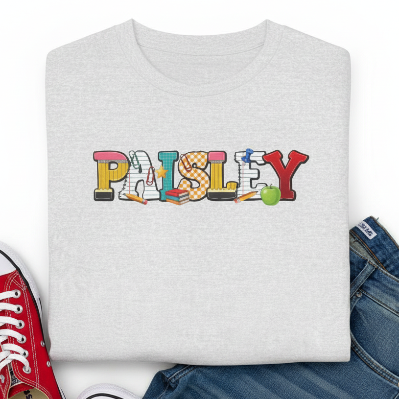 A folded light ash gray t-shirt featuring a colorful 'PAISLEY' graphic with school-themed icons, styled in a flat lay with blue jeans and a red sneaker on a white background.