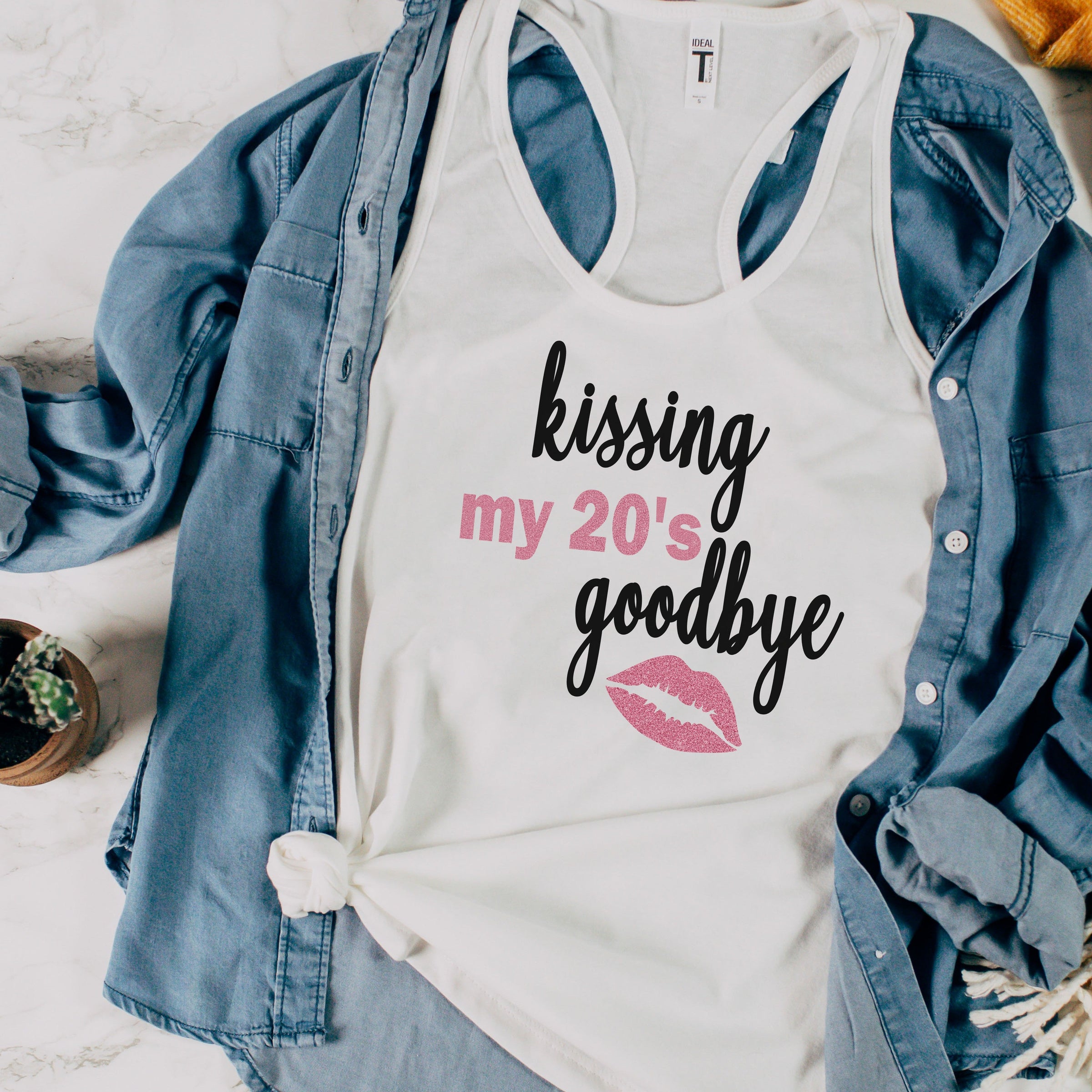 Womens white racerback tank on top of denim shirt. White tank says kissing my 20s goodbye. Kissing and goodbye are in black script. My 20s and a graphic of lips in pink glitter with my 20s written in block font.
