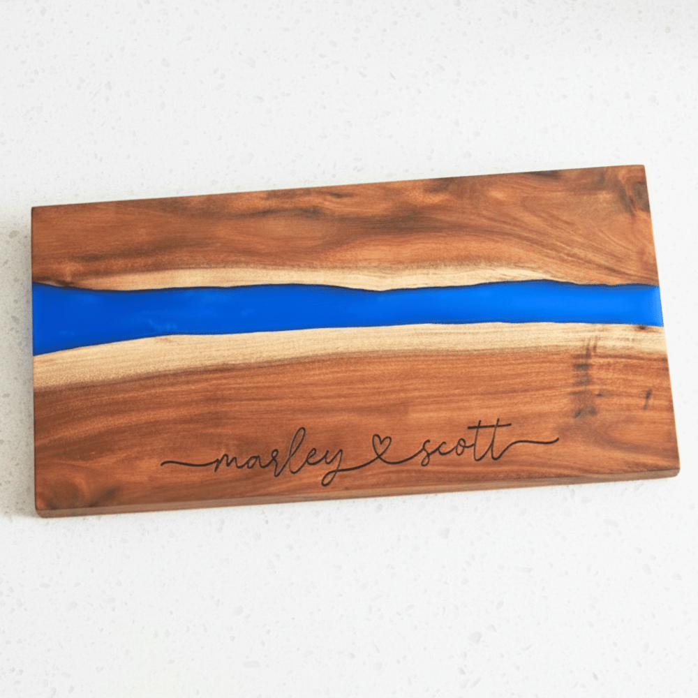 Wooden board with blue resin inlay and engraved names connected with a heart
 on a white background