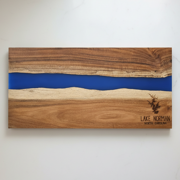 Acacia wood charcuterie board with blue resin inlay and 'Lake Norman North Carolina' text engraved in bottom right corner laying on a white kitchen counter