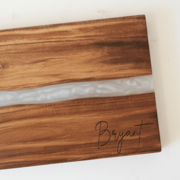 close up of wooden cutting board with 'Bryant' engraving in the bottom right corner and a white resin inlay through middle of board sitting on a white background