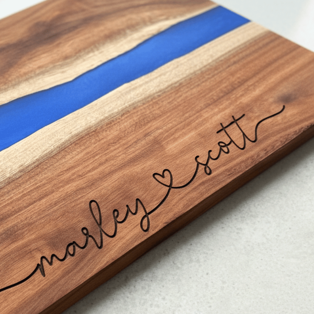 Wooden cutting board with blue resin strip and engraved names in a modern script font connected with a heart on a light background