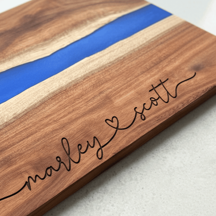 Wooden cutting board with blue resin strip and engraved names in a modern script font connected with a heart on a light background