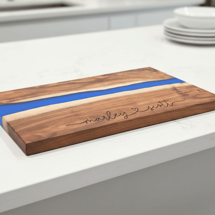 Wooden cutting board with blue resin inlay and engraved text of 2 script names connected with a heart in the middle on a kitchen counter.