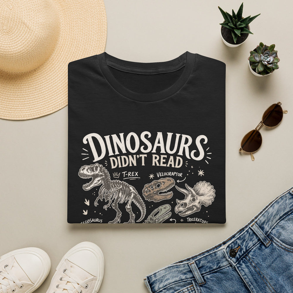 A folded black graphic T-shirt is displayed in a styled flat lay on a light beige surface. The shirt features white text reading 'DINOSAURS DIDN'T READ' with dinosaur illustrations below.