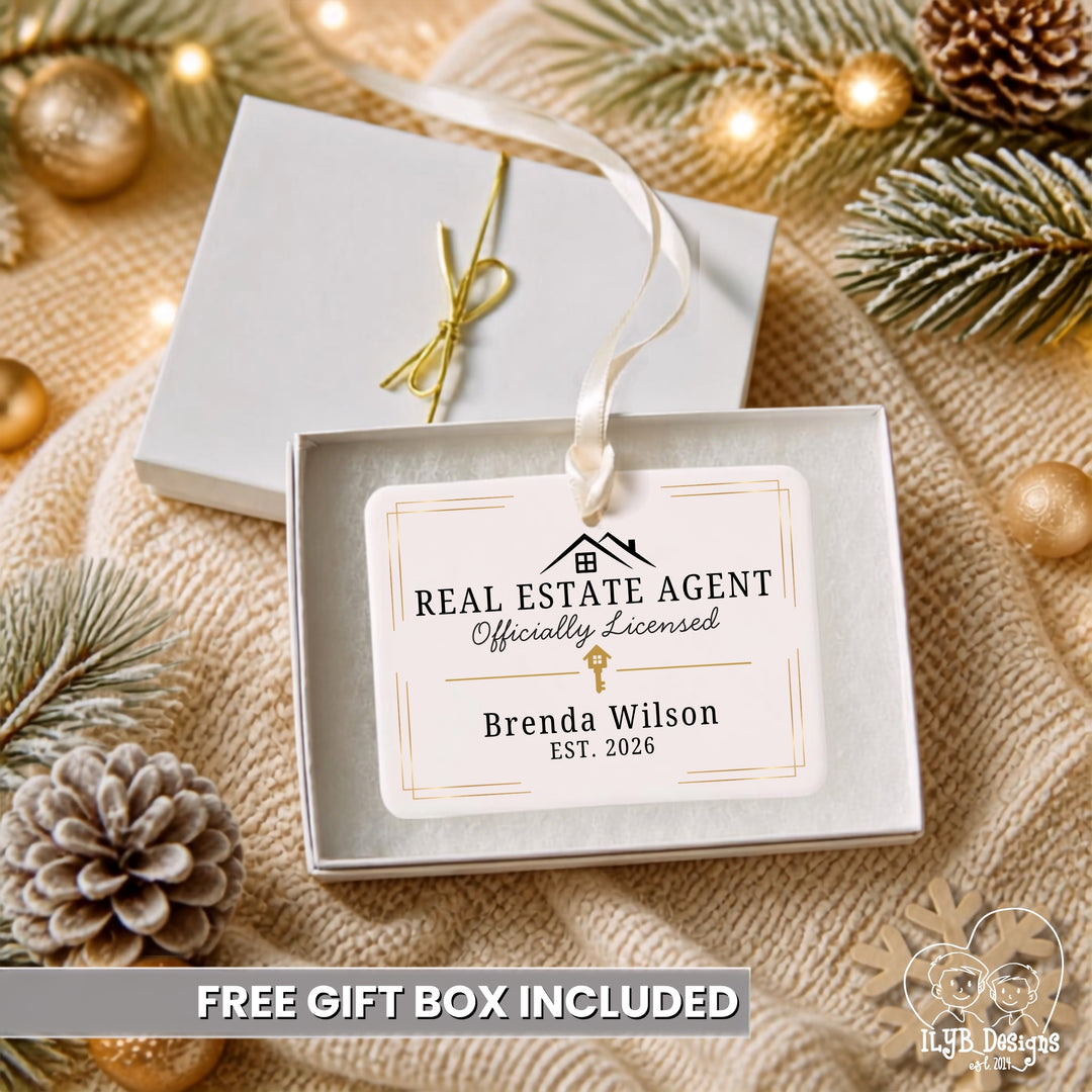 A personalized real estate agent rectangle shaped ornament hanging from a white ribbon in an open white gift box, surrounded by holiday greenery, pinecones, and gold ornaments on a beige knit blanket. The ornament reads &