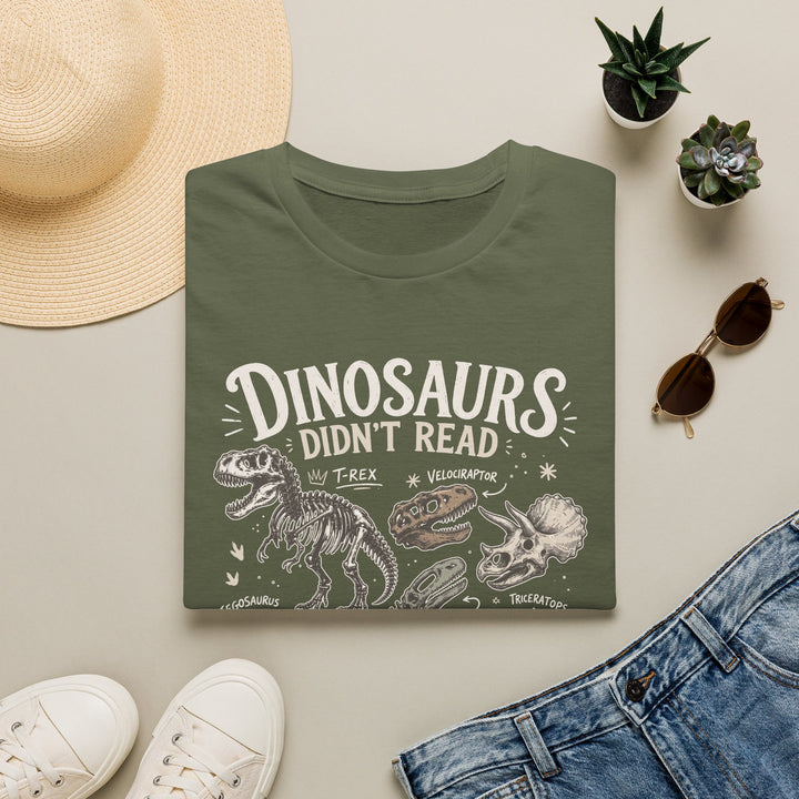 Dark gray t-shirt with 'DINOSAURS DIDN'T READ AND WE KNOW WHAT HAPPENED TO THEM' design on a light surface with accessories like a hat, sunglasses, and shoes.