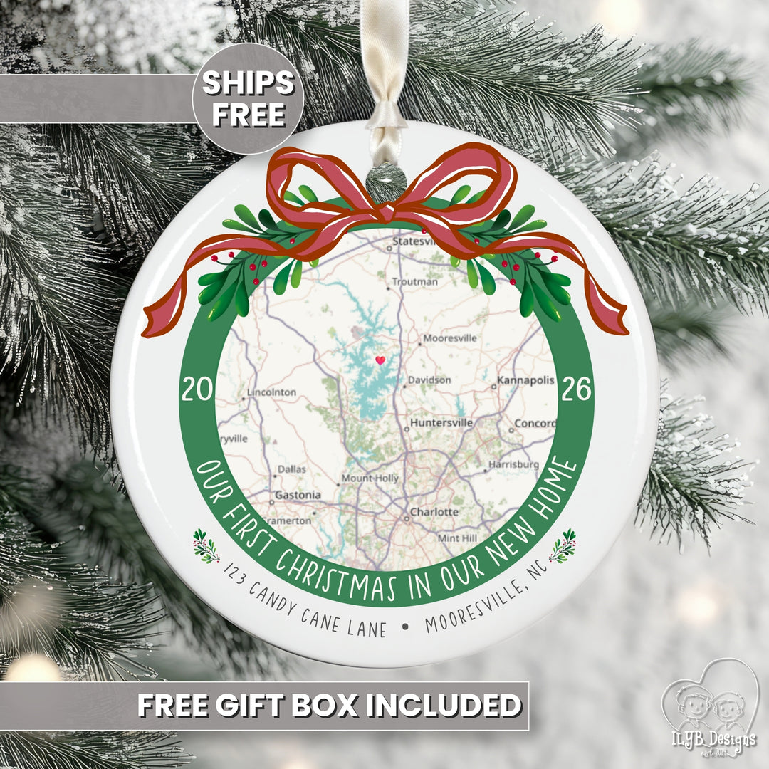 A round white Christmas ornament featuring a printed map with a red ribbon and green wreath design, hanging against snowy pine branches. Text on the ornament reads 'Our First Christmas in Our New Home' with a street address.