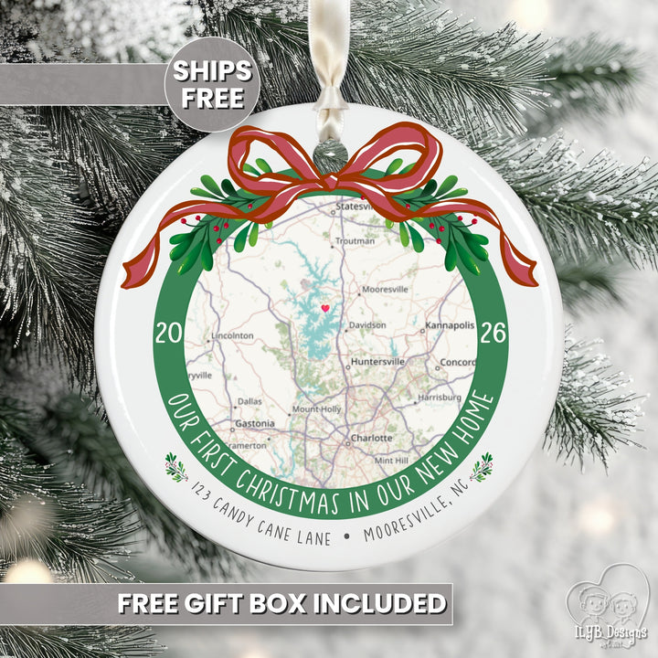 A round white Christmas ornament featuring a printed map with a red ribbon and green wreath design, hanging against snowy pine branches. Text on the ornament reads &
