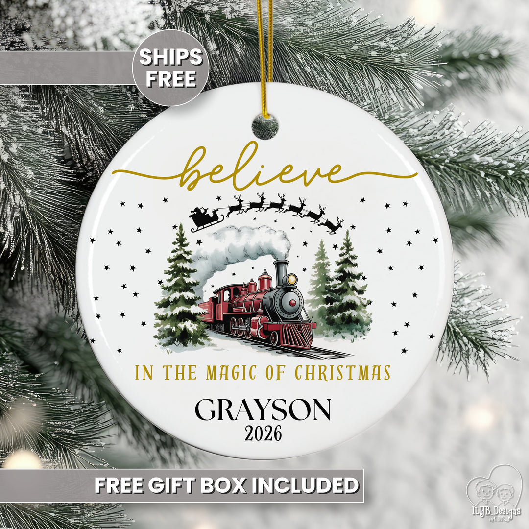 Personalized ceramic train Christmas ornament reading "believe in the magic of Christmas" with a red steam engine, Santa's sleigh, and a name and year, hanging on a frosted pine branch.
