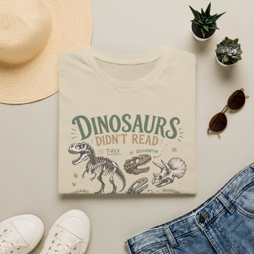 A folded cream-colored graphic T-shirt is styled in a flat lay on a light beige surface. The shirt features a dinosaur-themed print reading 'DINOSAURS DIDN'T READ and we know what happened to them' with illustrations of a T-rex, skull, and Triceratops.