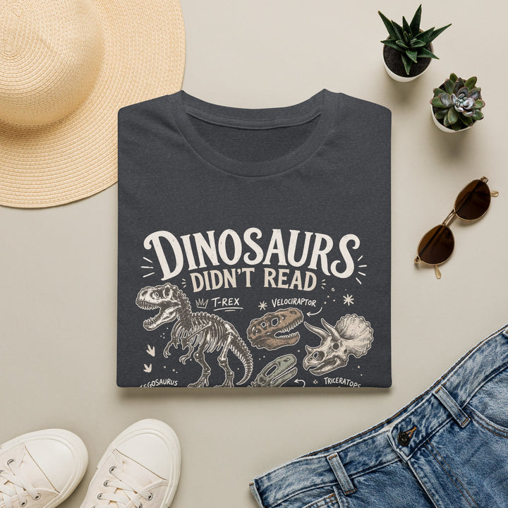 Dark heather gray t-shirt with 'dinosaurs didn't read and we know what happened to them' text, surrounded by a hat, sunglasses, shoes, and plants on a light surface.